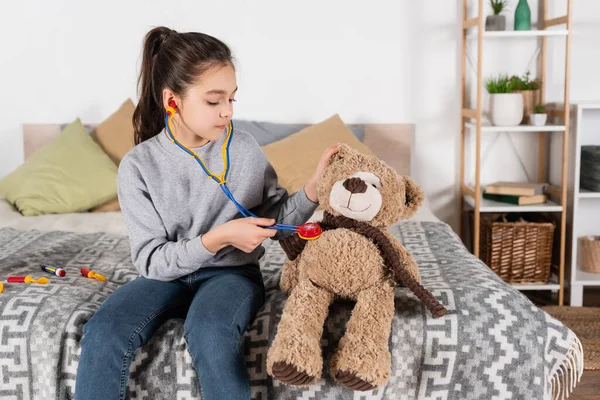 Morena preadolescente chica jugando en casa y examinando osito de peluche con estetoscopio de juguete - foto de stock