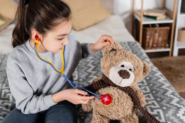Niña preadolescente examinando oso de peluche con estetoscopio de juguete mientras juega en casa - foto de stock