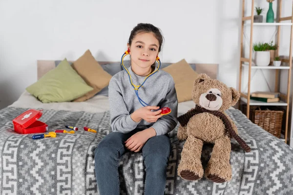 Дівчина з іграшковим стетоскопом дивиться на камеру біля плюшевого ведмедя і медичного набору — стокове фото