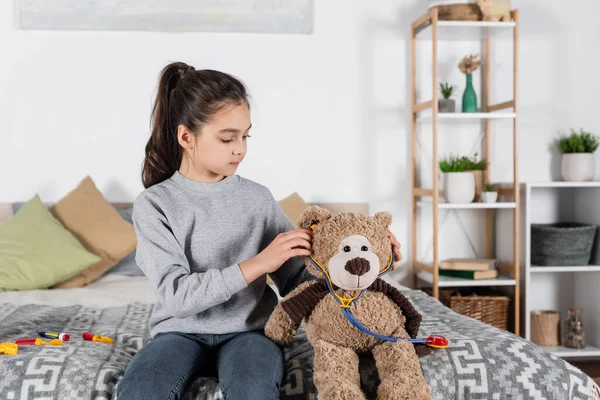 Дівчина кладе іграшковий стетоскоп на плюшевого ведмедя, граючи в спальні — стокове фото