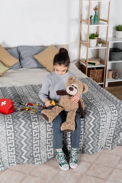 Високий кут зору дівчини, яка вивчає плюшевого ведмедя з неврологічним мальлюсом під час гри на ліжку — стокове фото