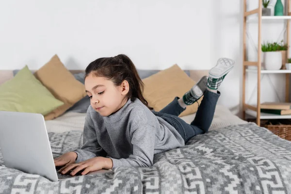 Visão de comprimento total da menina pré-adolescente deitada na cama e digitando no laptop — Fotografia de Stock