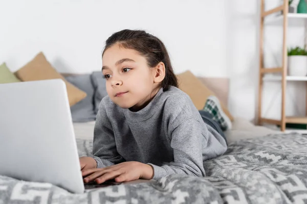 Preadolescente chica acostada en la cama y escribiendo en el ordenador portátil - foto de stock