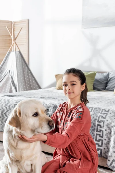 Preadolescente morena chica sonriendo a cámara cerca amarillo labrador en dormitorio - foto de stock