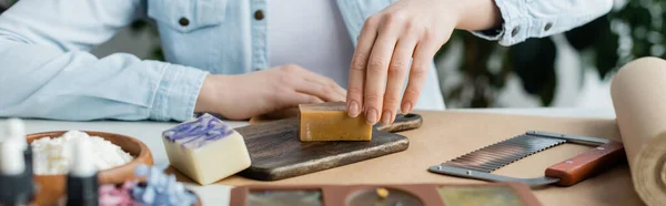 Vista recortada de la artesana haciendo jabón en la tabla de cortar cerca de suministros, pancarta - foto de stock