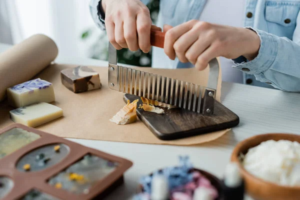 Cropped view of craftswoman cutting soap on board near blurred flowers at home — Stock Photo