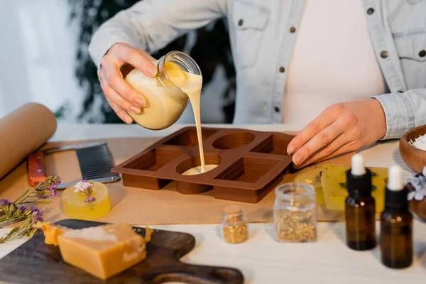 Cropped view of craftswoman pouring soap in silicone mold near flowers and essential oils on table — Stock Photo