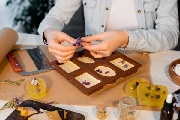 Vista ritagliata di artigiana che tiene fiori secchi vicino al sapone in stampo in silicone e forniture sul tavolo — Foto stock