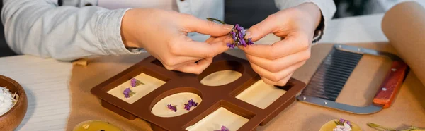 Vista recortada de la artesana sosteniendo flores cerca del jabón hecho a mano en molde de silicona en casa, pancarta - foto de stock