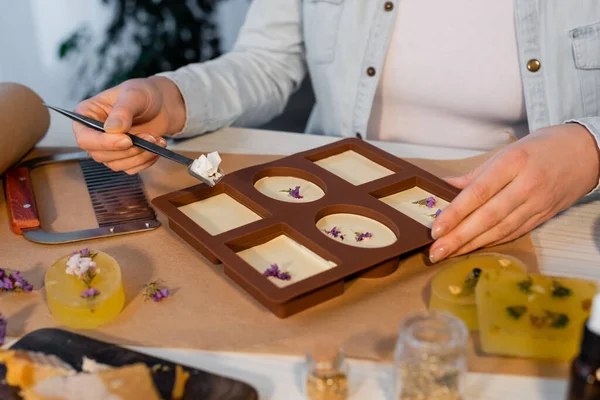 Ausgeschnittene Ansicht einer Handwerkerin, die Seifenflocken in der Nähe von Silikonform und Blumen auf dem Tisch hält — Stockfoto