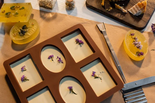 Vista superior de jabón hecho a mano con flores en molde de silicona cerca del cortador y papel artesanal en la mesa - foto de stock