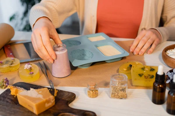 Ausgeschnittene Ansicht einer Handwerkerin, die ein Glas mit Seife in der Nähe von Silikonschimmel, ätherischen Ölen und Blumen auf den Tisch nimmt — Stockfoto