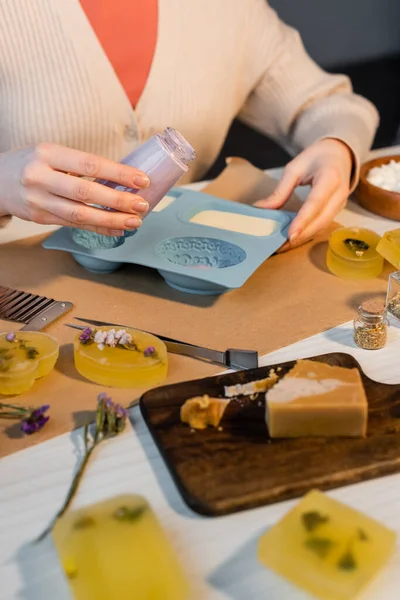 Vue recadrée de l'artisan tenant un bocal avec du savon liquide près de la moisissure de silicone et des fleurs sèches sur la table — Photo de stock