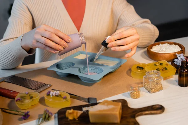 Vue recadrée de l'artisan versant du savon dans un moule près des fleurs, coupeur et flocons sur la table — Photo de stock