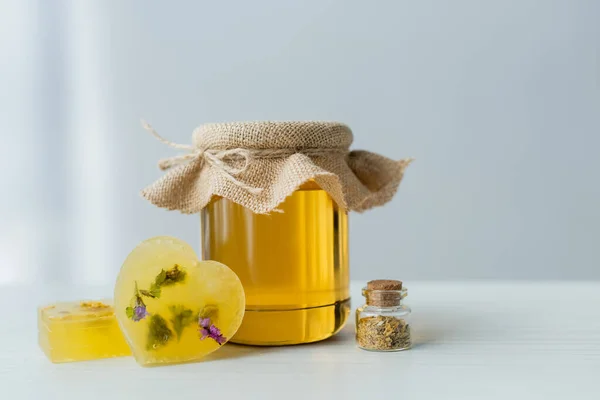 Jar with honey near handmade soap bars and herbs on table on grey background — Stock Photo