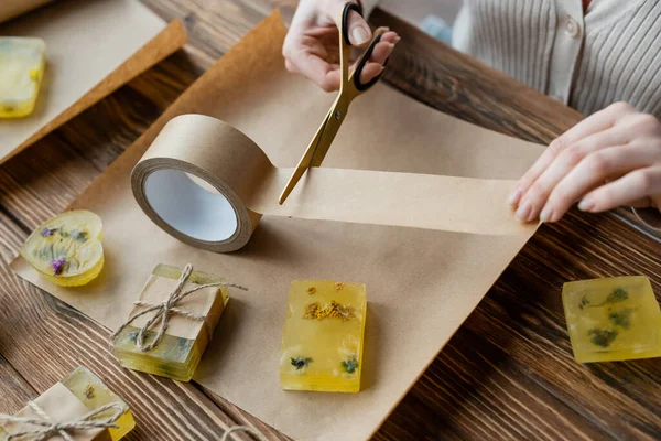 Vista recortada de la artesana cortando cinta adhesiva cerca de jabón hecho a mano en papel artesanal - foto de stock