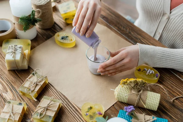 Vue du dessus de l'artisan versant de la cire liquide dans du verre près des barres de savon et du papier artisanal — Photo de stock