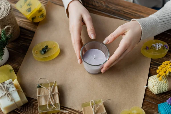 Vue du dessus de l'artisan tenant une bougie faite à la main en verre près des barres de savon sur du papier artisanal — Photo de stock