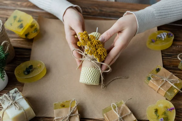 Vue du dessus de l'artisan tenant une bougie faite à la main avec des fleurs près de barres de savon sur du papier artisanal — Photo de stock