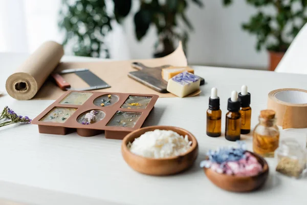 Jabón hecho a mano en molde de silicona cerca de aceites esenciales y flores borrosas en la mesa - foto de stock