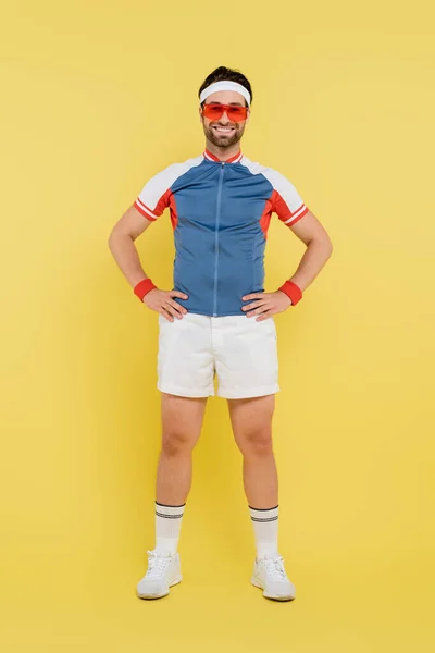 Полная длина улыбающегося спортсмена в солнечных очках, держащегося за руки на бёдрах на жёлтом фоне — стоковое фото