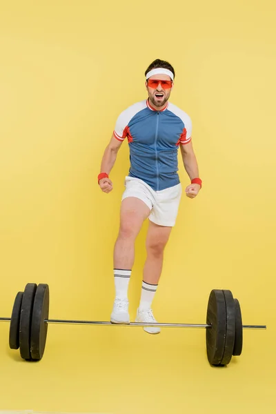 Jovem desportista em óculos de sol mostrando músculos perto barbell no fundo amarelo — Fotografia de Stock