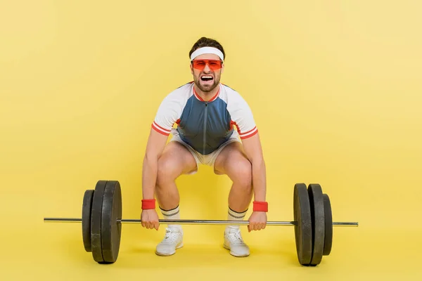 Sportsman screaming while lifting barbell on yellow background — Stock Photo