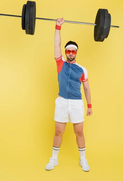 Sportler mit Sonnenbrille hebt die Langhantel und blickt in die Kamera auf gelbem Hintergrund — Stockfoto