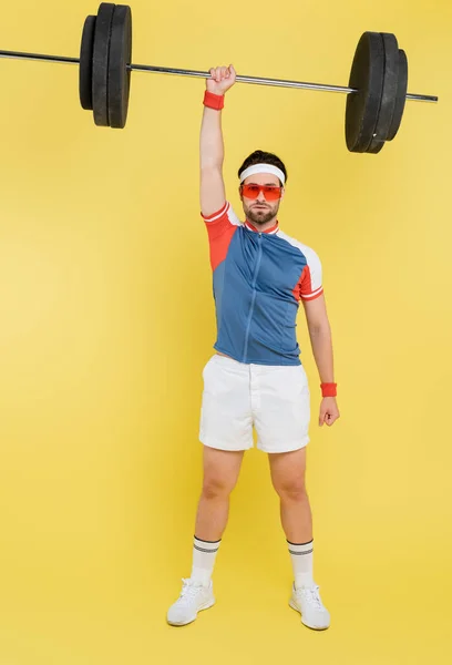 Sportsman en lunettes de soleil levant haltère et regardant la caméra sur fond jaune — Photo de stock