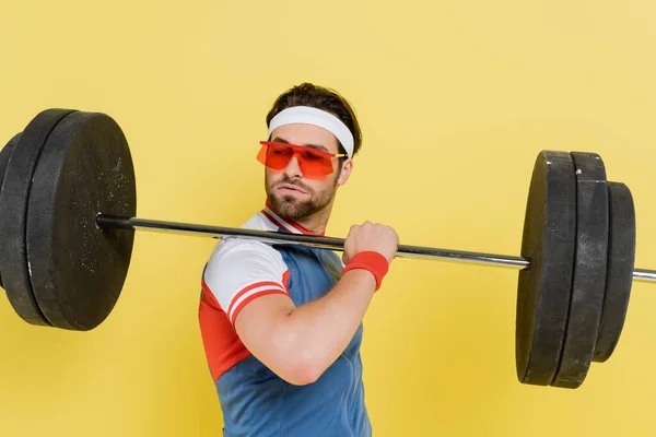 Young sportsman in sunglasses holding barbell isolated on yellow — Stock Photo