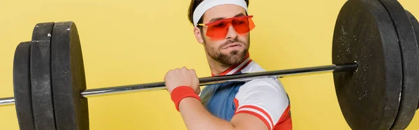 Sportler mit Sonnenbrille blickt auf Langhantel isoliert auf gelbem Banner — Stockfoto