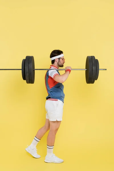 Side view of sportsman in sunglasses holding barbell on shoulder on yellow background — Stock Photo