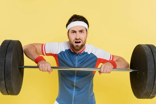 Beleidigter Sportler blickt beim Heben der Langhantel isoliert auf gelb in die Kamera — Stockfoto