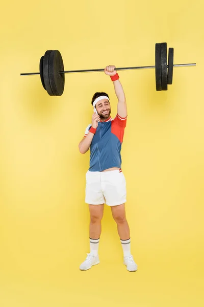 Comprimento total do esportista sorrindo falando no celular e levantando a barra no fundo amarelo — Fotografia de Stock