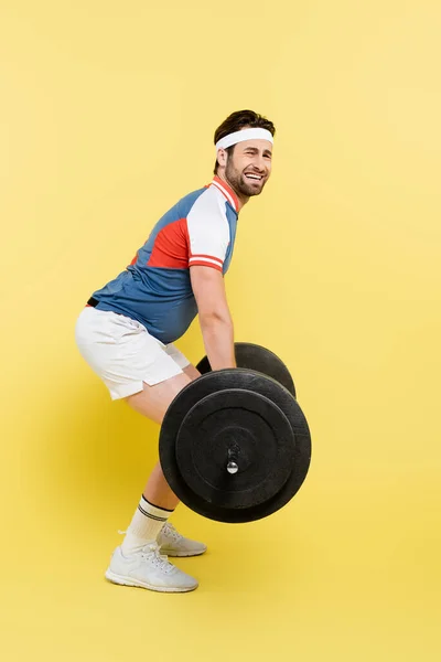 Full length of young sportsman suffering while lifting barbell on yellow background — Stock Photo