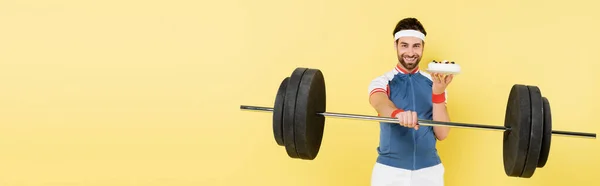 Positive sportsman holding tasty cake and barbell isolated on yellow, banner — Stock Photo