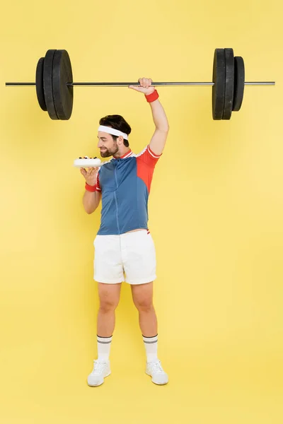 Side view of young sportsman smelling cake and lifting barbell on yellow background — Stock Photo