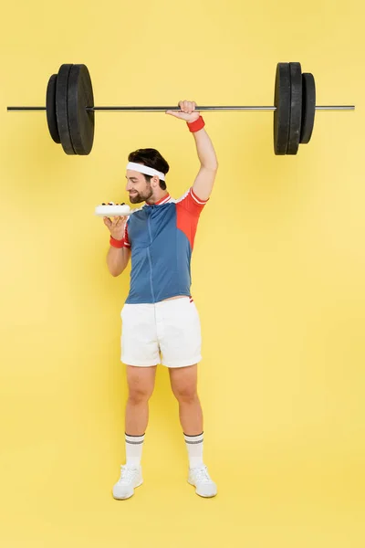 Vue latérale du jeune sportif sentant le gâteau et soulevant l'haltère sur fond jaune — Photo de stock