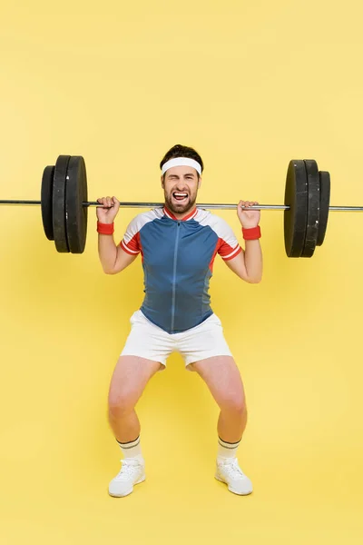 Молодой спортсмен гримасит, поднимая штангу на желтый фон — стоковое фото