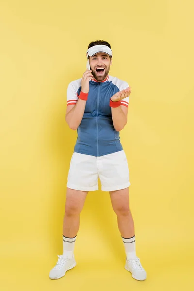 Longitud completa de deportista emocionado hablando en el teléfono inteligente y mirando a la cámara en el fondo amarillo - foto de stock