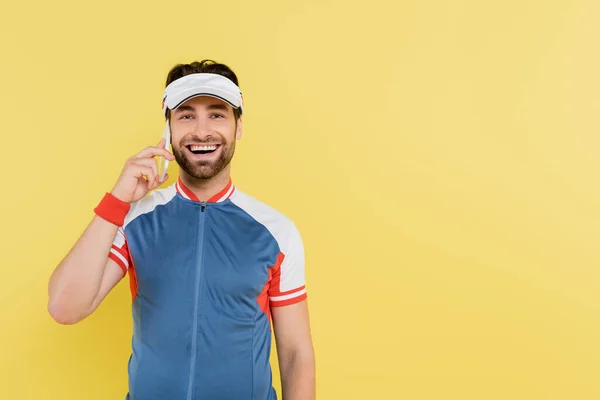 Uomo positivo in abbigliamento sportivo e visiera che parla sul cellulare isolato su giallo — Foto stock