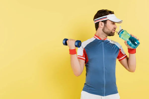 Vue latérale du sportif tenant une bouteille de sport et un haltère isolés sur jaune — Photo de stock