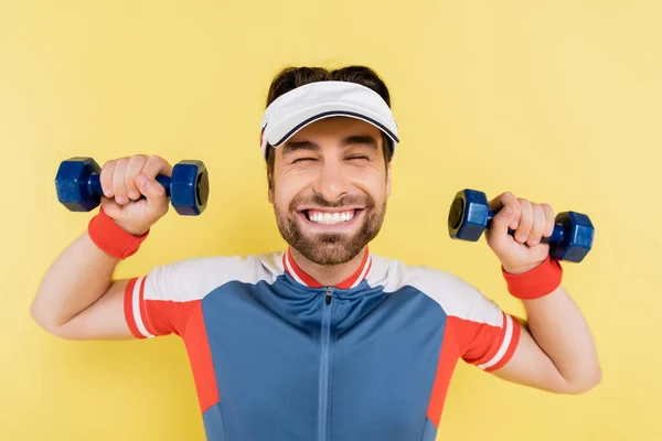 Cheerful sportsman in visor holding dumbbells isolated on yellow — Stock Photo