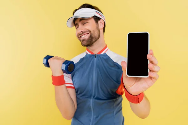 Treinamento esportista desfocado com haltere e segurando celular com tela em branco isolada no amarelo — Fotografia de Stock