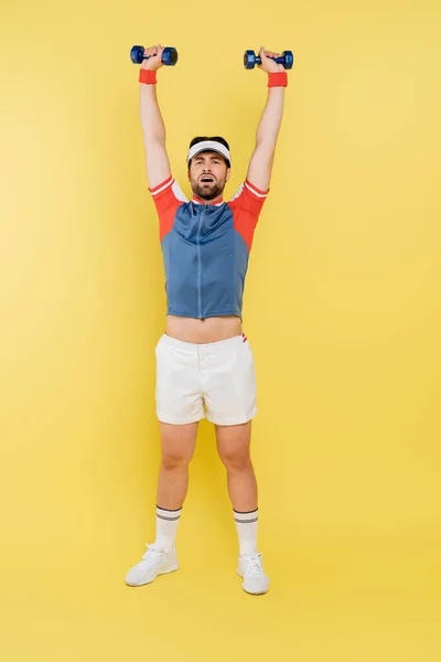 Full length of tired sportsman training with dumbbells on yellow background — Stock Photo