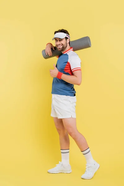 Longitud completa de deportista sonriente mostrando como y sosteniendo alfombra de fitness sobre fondo amarillo - foto de stock