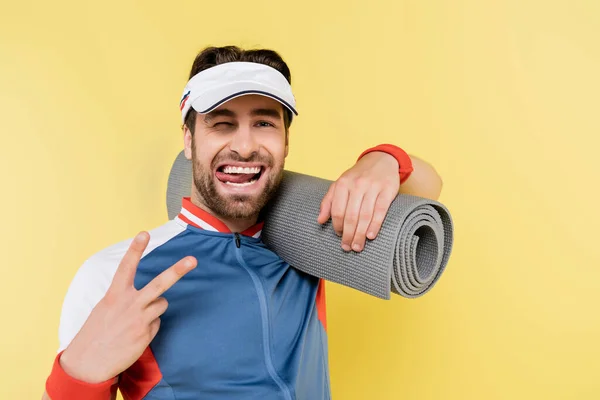 Esportista em viseira segurando tapete fitness e mostrando sinal de paz isolado no amarelo — Fotografia de Stock