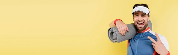 Joven deportista mostrando un signo de paz y sosteniendo una alfombra de fitness aislada en amarillo, pancarta - foto de stock