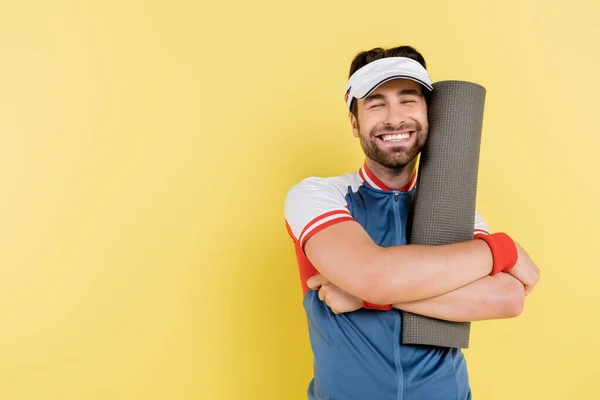 Esportista alegre em viseira abraçando tapete fitness isolado em amarelo — Fotografia de Stock