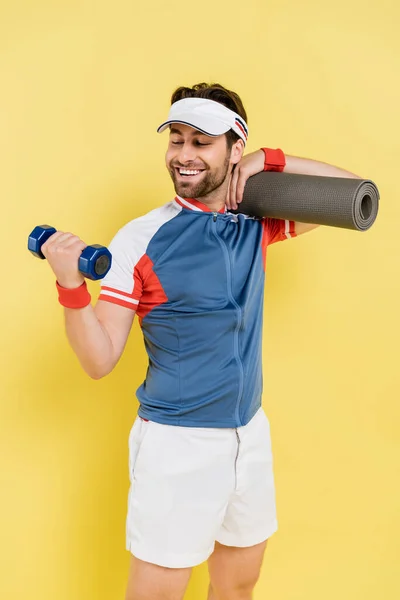 Sportsman sorrindo segurando tapete de fitness e treinamento com halteres isolados em amarelo — Fotografia de Stock